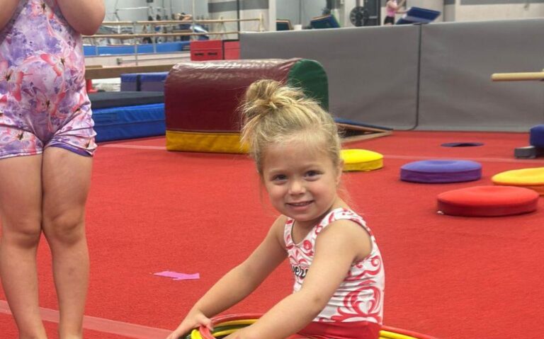 Preschool gymnast sitting on a red carpet.